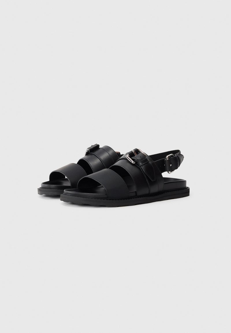 Black sandals featuring multiple wide straps, adjustable buckle, flat sole, and a smooth texture. Minimalist design with no prominent patterns.