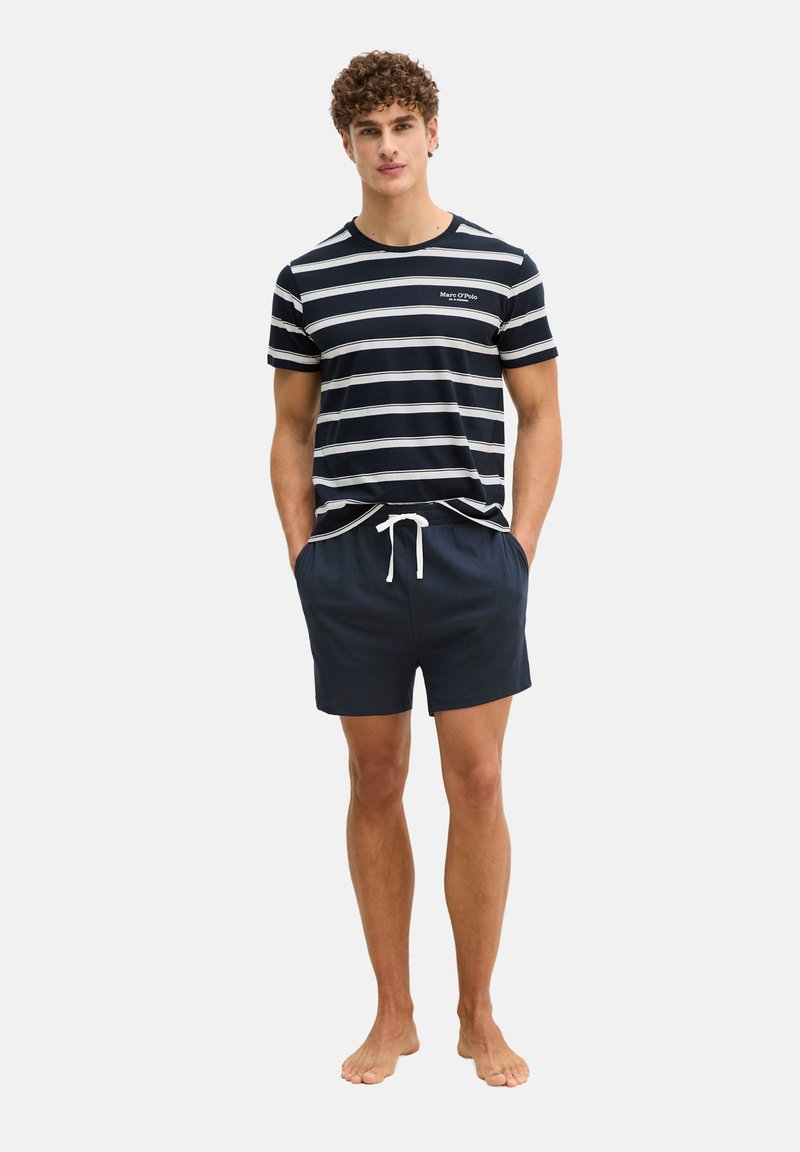 Jonge man staande op blote voeten, gekleed in een marineblauw-wit gestreept T-shirt en marineblauwe korte broek met een wit trekkoord, handen in de zakken.