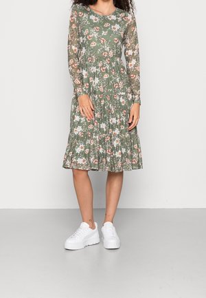 Nainen pukeutuneena vihreään kukalliseen pitkähihainen midimekkoon, yhdistettynä valkoisiin korokepohjakenkiin, seisoo yksinkertaista vaaleaa seinää vasten.