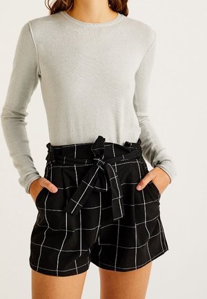 Femme portant un haut à manches longues gris clair et un short taille haute noir avec un motif en grille blanche et une ceinture nouée, les mains dans les poches.