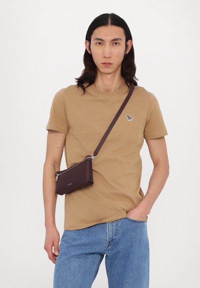 Tan cotton t-shirt with short sleeves, paired with denim jeans. Crossbody bag in dark burgundy leather with silver zipper and logo detail.