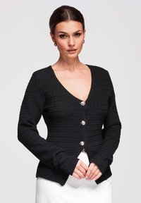 Femme aux cheveux foncés coiffés en chignon bas, portant un cardigan noir texturé boutonné et une jupe blanche, debout les mains jointes.