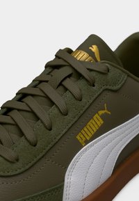 Αθλητικό παπούτσι σε ελαιί πράσινο με ανώτερο μέρος από σουέντ και συνθετικό δέρμα, λευκή ενδιάμεση σόλα με υφή κυματοειδούς σχεδίου, χρυσές λεπτομέρειες λογότυπου και επίπεδες κορδόνια.