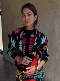 Pletený čierny cardigan s farebnými kvetinovými a geometrickými vzormi, so zapínaním na gombíky a rebrovanými manžetami. Zlaté prstene a náramky ako akcenty.