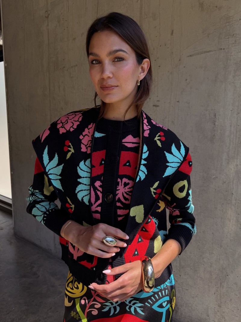 Pletený čierny cardigan s farebnými kvetinovými a geometrickými vzormi, so zapínaním na gombíky a rebrovanými manžetami. Zlaté prstene a náramky ako akcenty.