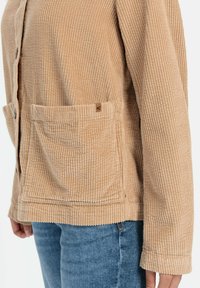 Kordjacke in Beige, mit Knopfverschluss, Fronttaschen, gerippter Textur und langen Ärmeln. Kombiniert mit blauer Jeans.