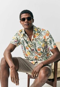 Kortærmet skjorte lavet af letvægtsstof med et farverigt mønster af frugter og blade. Modellen bærer solbriller og beige shorts og sidder ned.