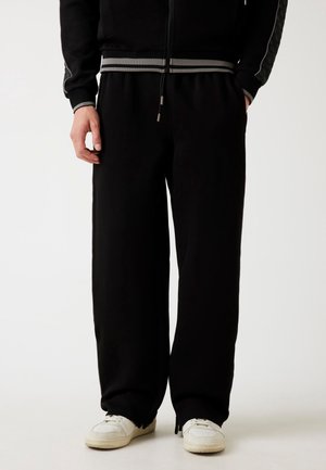 Person, die schwarze Hosen mit weitem Bein, schwarze Jacke und weiße Sneakers trägt, steht vor einem schlichten Hintergrund.