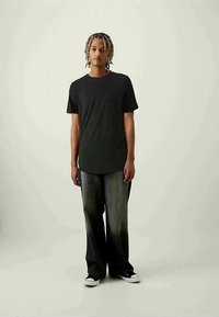 Jovem com dreadlocks loiros, a usar uma t-shirt preta, jeans largos desbotados pretos e ténis preto e branco, a pôr-se de pé contra um fundo simples.