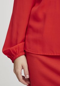 Rote Bluse mit langen, gerafften Ärmeln und einem lässigen Schnitt. Glatte Textur und ein einfacher Saumabschluss. Hand, die gegen den Stoff ruht.