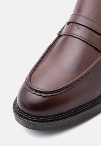 Gros plan sur un mocassin en cuir marron avec des coutures détaillées et une semelle en caoutchouc noire sur un fond blanc.