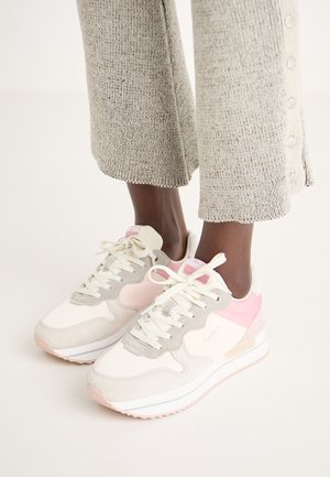 Person wearing beige knit pants and white sneakers with pink and gray accents, standing on a light background.