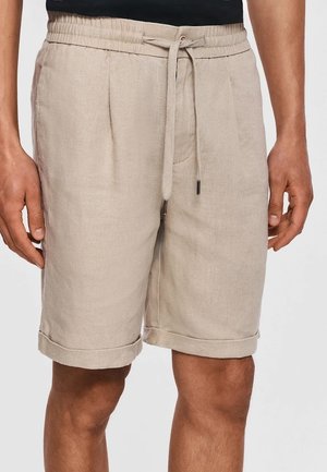 Beżowe krótkie spodenki do kolan z podwiniętymi brzegami, plisami z przodu, bocznymi kieszeniami i sznurkiem w talii, noszone przez osobę w czarnej koszuli.