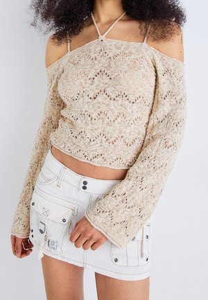 Kobieta ubrana w beżowy, ażurowy sweter z odsłoniętymi ramionami i długimi rękawami oraz białą mini spódnicę z dużymi kieszeniami i guzikami.