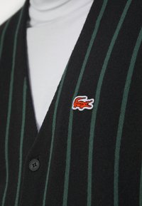 Black cardigan with vertical green stripes, featuring a round black button and a small red and white crocodile logo on the chest.