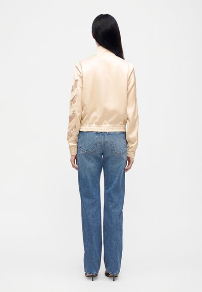 Femme aux longs cheveux noirs portant une veste bomber en satin beige et un jean bleu droit taille haute, debout de dos sur fond blanc.