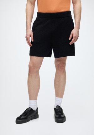 Mann trägt schwarze strukturierte Shorts, weiße Kniestrümpfe und schwarze niedrige Sneakers, die Hände entspannt seitlich am Körper vor weißem Hintergrund.