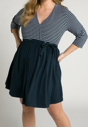 Woman wearing a black and white striped wrap top dress with three-quarter sleeves and a tied waist belt, standing with hands on hips.