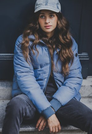 Blauer Pufferjacke mit hohem Kragen, Reißverschluss und gerippten Bündchen. Kombiniert mit dunklen Jeans und einer Denim-Kappe, sitzt auf Steinstufen.