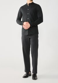 Camicia nera a maniche lunghe con colletto alto, bottoni bianchi e una tasca, abbinata a pantaloni scuri e scarpe nere.