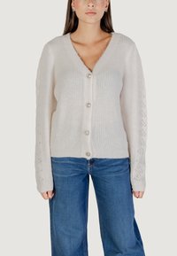 Cárdigan crema de pico con textura acanalada, seis botones decorativos y un patrón de encaje en las mangas. Combinado con jeans de pierna ancha en azul.