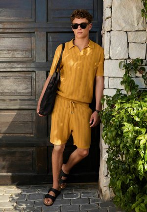 Junger Mann in ockerfarbenen Strickshorts und Poloshirt, schwarze Sandalen und Sonnenbrille, der sich an einer Steinmauer neben grünen Pflanzen lehnt.