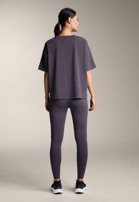 Žena stojí chrbtom otočená, oblečená v voľnom tmavosivom tričku, zodpovedajúcich legínach a čiernych teniskách s bielymi podrážkami na béžovom pozadí.