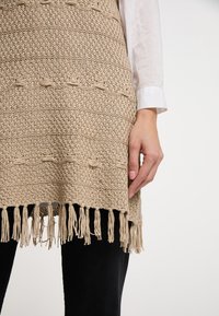 Indumento a maglia beige con un motivo testurizzato e dettagli annodati. Orlo con frange, indossato sopra pantaloni neri, con maniche lunghe sotto.