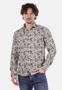 Camicia a maniche lunghe color crema con un intricato motivo floreale nero, colletto e polsini con bottoni. Abbinata a jeans blu.