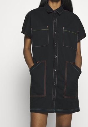 Czarna sukienka na guziki z krótkim rękawem, z czterema dużymi kieszeniami i kolorowymi kontrastowymi przeszyciami, noszona przez osobę z rękami w kieszeniach.