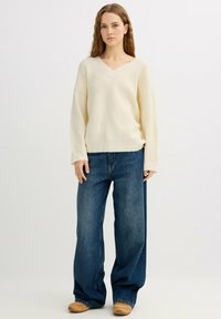 Suéter de punto color crema con cuello en V, hombros caídos y textura acanalada, combinado con unos vaqueros de mezclilla azul oscuro de corte holgado.