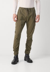 Olivgrüne Cargo-Hosen mit elastischem Bund und Kordelzug. Mit seitlichen Taschen und schmal zulaufenden Säumen. Kombiniert mit schwarzen Knöchelstiefeln.