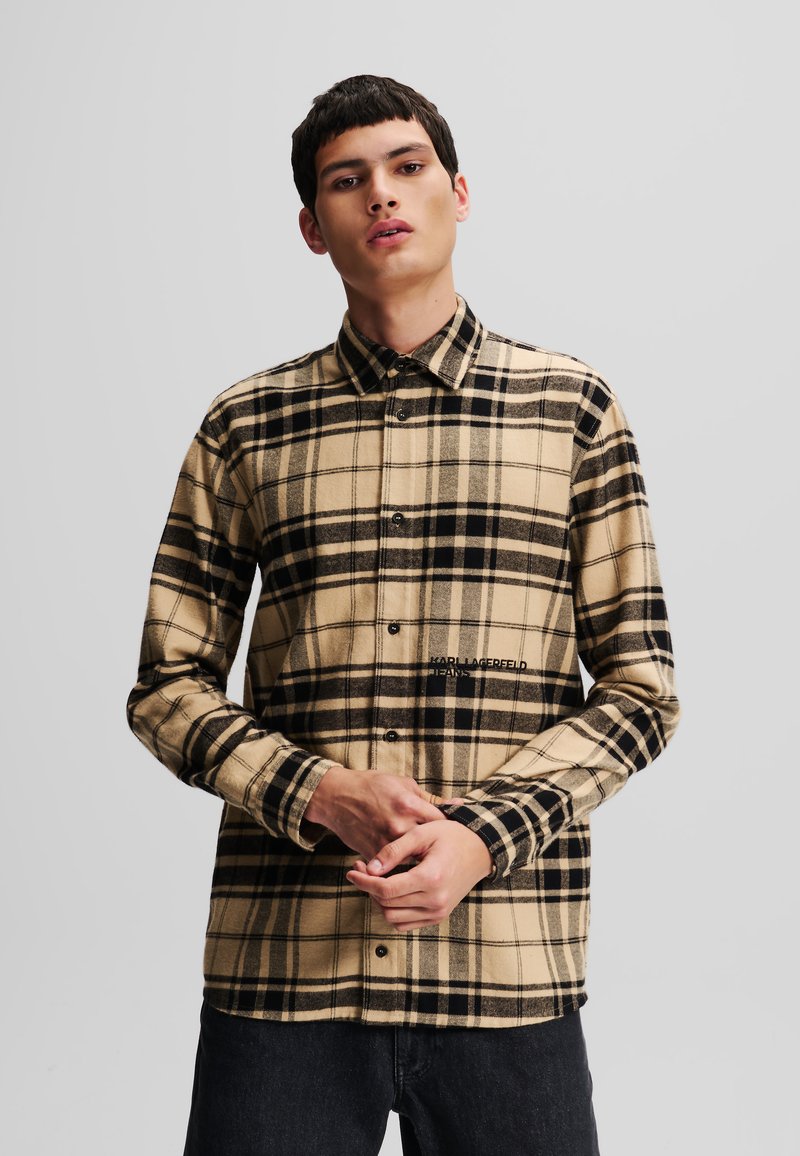 Camisa de franela de cuadros beige y negro con cuello abotonado, cierre frontal con botones y logo de la marca en el lado izquierdo. Mangas largas.
