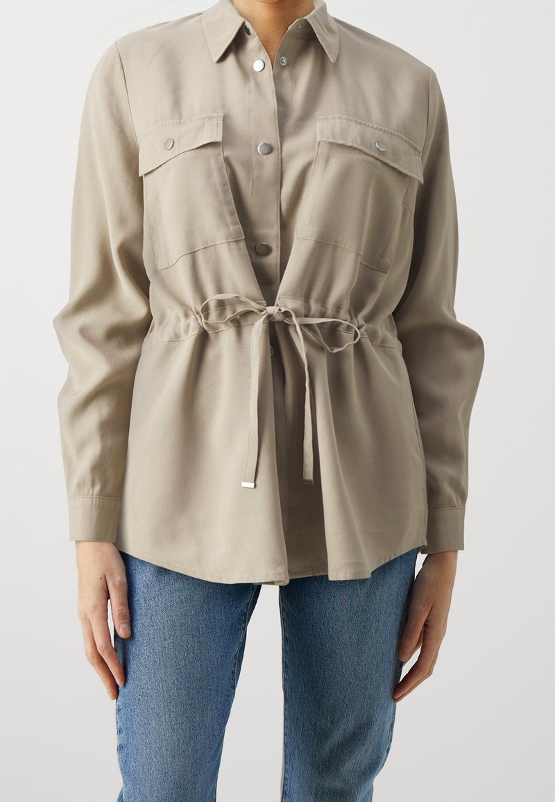 Chemise beige à manches longues avec poches poitrine et boutons sur le devant, nouée à la taille, portée avec un jean bleu sur une personne debout devant un fond uni.