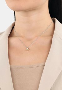 Silverhalsband med en liten guldberlock i form av en anka; bärs av en person med en beige topp och en ljusblå blazer.