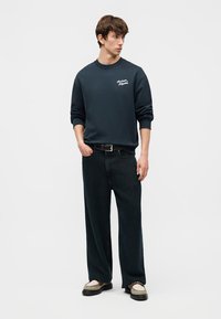 Νεαρός άνδρας που φοράει ναυτικό μπλε φούτερ με λευκό κείμενο, φαρδιά μαύρα τζιν, μπεζ μοκασίνια, στέκεται με το ένα χέρι στην τσέπη κοιτάζοντας πλάγια.