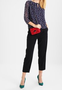 Marinebluse med et mønster af små, farverige former, parret med sorte cropped bukser. Modellen holder en rød clutch og bærer teal hæle.
