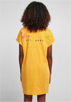 Person mit lockigem Haar trägt ein gelbes kurzärmeliges Kleid mit buntem Text und "EST. 2005" auf dem Rücken vor einem einfachen Hintergrund.