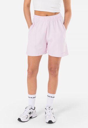 Personne portant un short rose clair à taille élastique, un débardeur court blanc, des chaussettes blanches et des baskets blanches New Balance, debout avec les mains dans les poches.