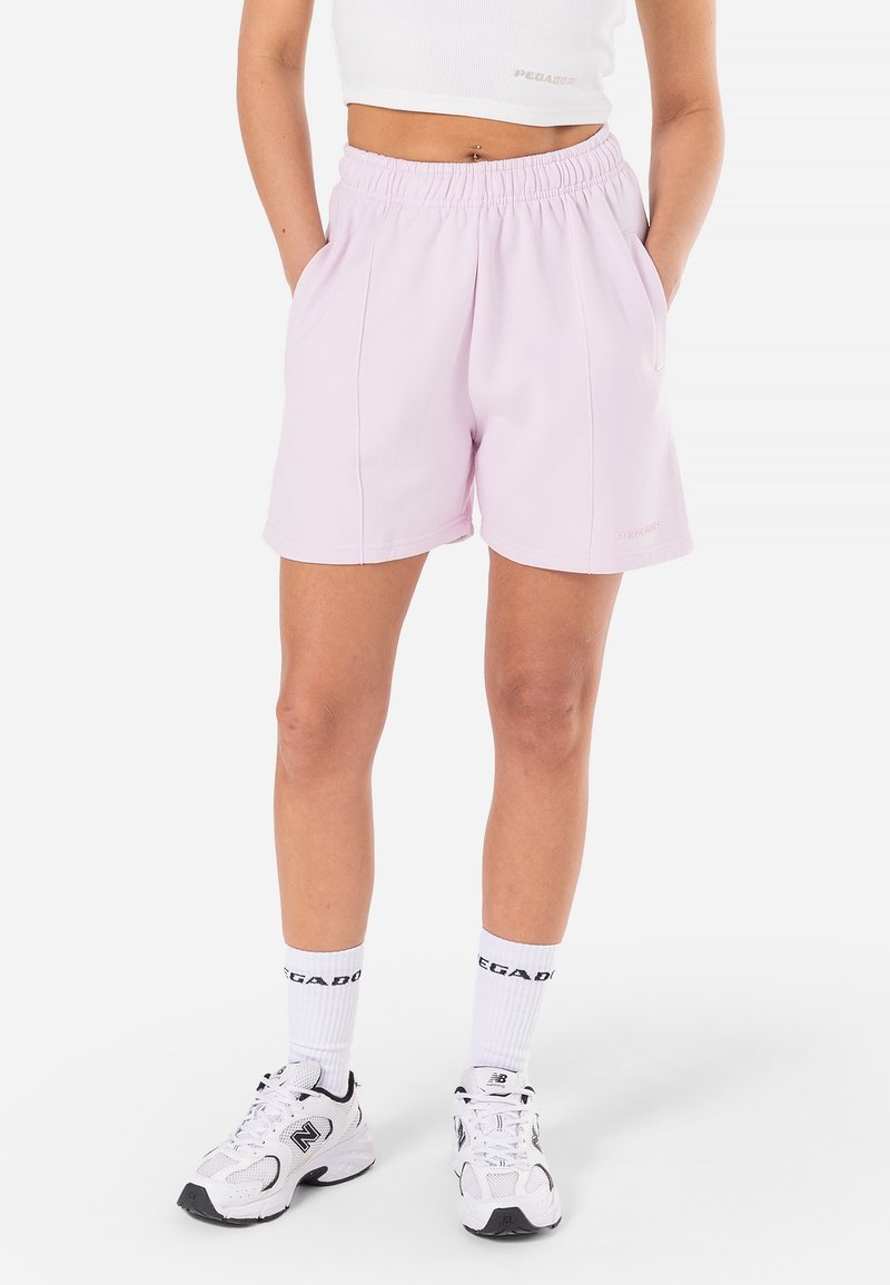 Personne portant un short rose clair à taille élastique, un débardeur court blanc, des chaussettes blanches et des baskets blanches New Balance, debout avec les mains dans les poches.