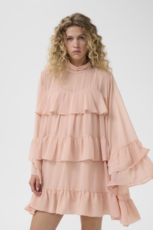 Γυναίκα με σγουρά ξανθά μαλλιά που φορεί ένα χαλαρό, στρωματοποιημένο ροδακινί φόρεμα με φαρδιά βολάν μανίκια, μπροστά σε απλό φωτεινό φόντο.