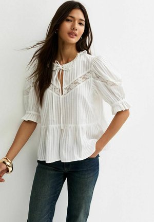 Kobieta o długich ciemnych włosach nosi białą bluzkę z panelami z koronki i bufkami oraz ciemnoniebieskie dżinsy, pozując na białym tle.