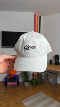 Hand holding a white Gant baseball cap inside a modern living room with plants, a TV, and colorful wall stripes.