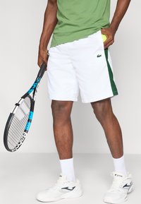 Λευκά σορτς τένις με πράσινες λωρίδες στο πλάι, σε συνδυασμό με πράσινο μπλουζάκι. Κρατά μια μπάλα τένις και μια μαύρη και μπλε ρακέτα τένις.