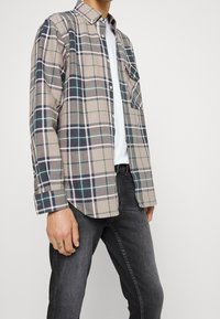 Camisa de cuadros beige con acentos en marino, verde y blanco, diseño abotonado, mangas largas y bolsillo en el pecho izquierdo, combinada con jeans oscuros.