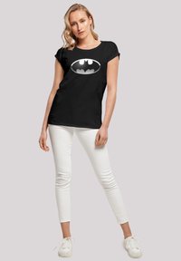 Svart t-skjorte med en rund Batman-logo i hvitt, med en teksturert design. Kombinert med hvite slim-fit bukser og hvite sneakers.