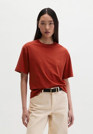 Jonge vrouw met lang, stijl zwart haar, een roestkleurig T-shirt met korte mouwen, ingestopt in een beige broek met hoge taille en zwarte riem.