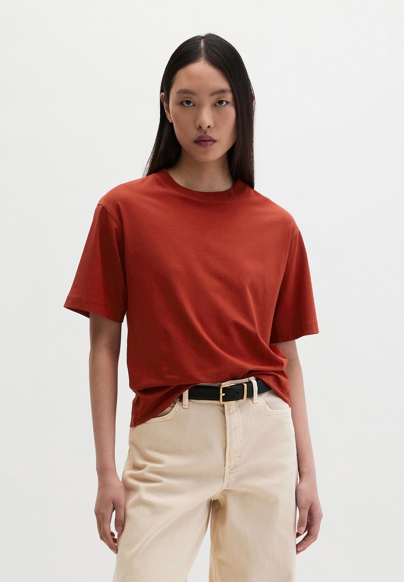 Jeune femme aux longs cheveux noirs et raides, portant un t-shirt à manches courtes couleur rouille, rentré dans un pantalon beige taille haute avec une ceinture noire.
