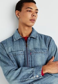Chaqueta de denim en azul claro con cierre de cremallera, dos bolsillos en el pecho y detalles de costura vertical. Llevada sobre una camisa roja, con puños abotonados.