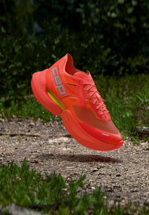 Bright orange running shoe with reflective CEP logo hovering above a gravel path surrounded by green grass and foliage.